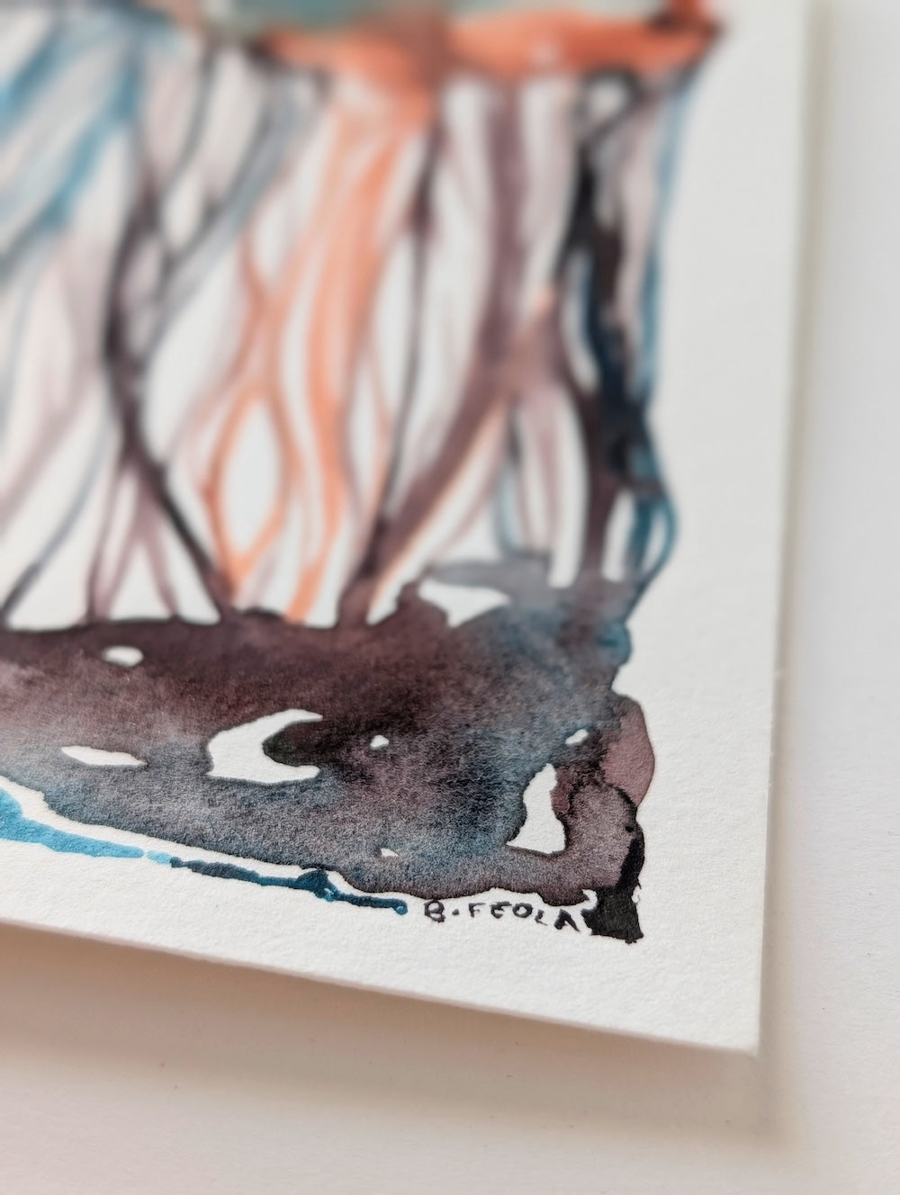 Close-up of an abstract watercolor painting on textured white paper, showing flowing orange, blue, black, and gray brushstrokes with a visible artist signature near the bottom.